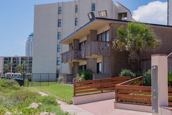 A multi-story building with balconies, a palm tree, and a fenced garden area.