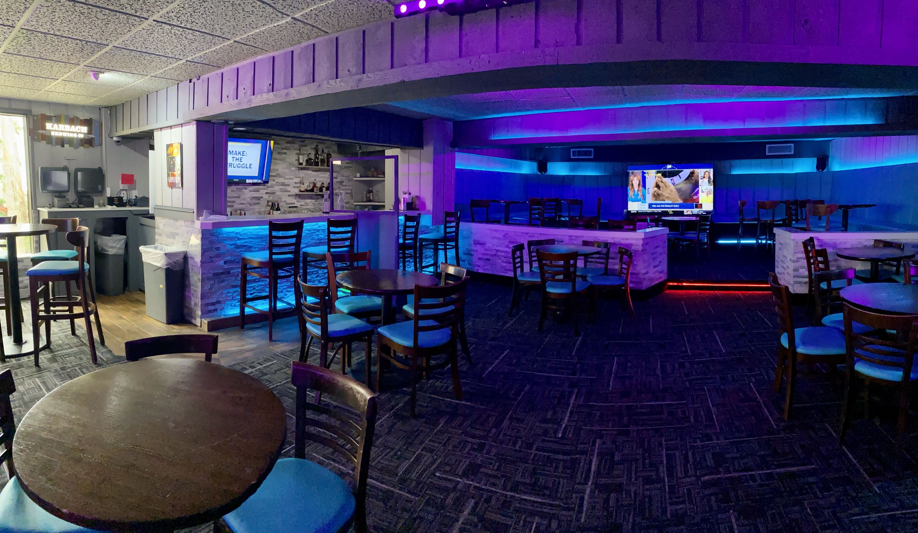 A bar area with tables and chairs, a counter with a menu board, and a television screen displaying a program.