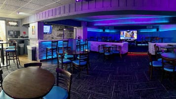 A bar area with tables and chairs, a counter with a menu board, and a television screen displaying a program.