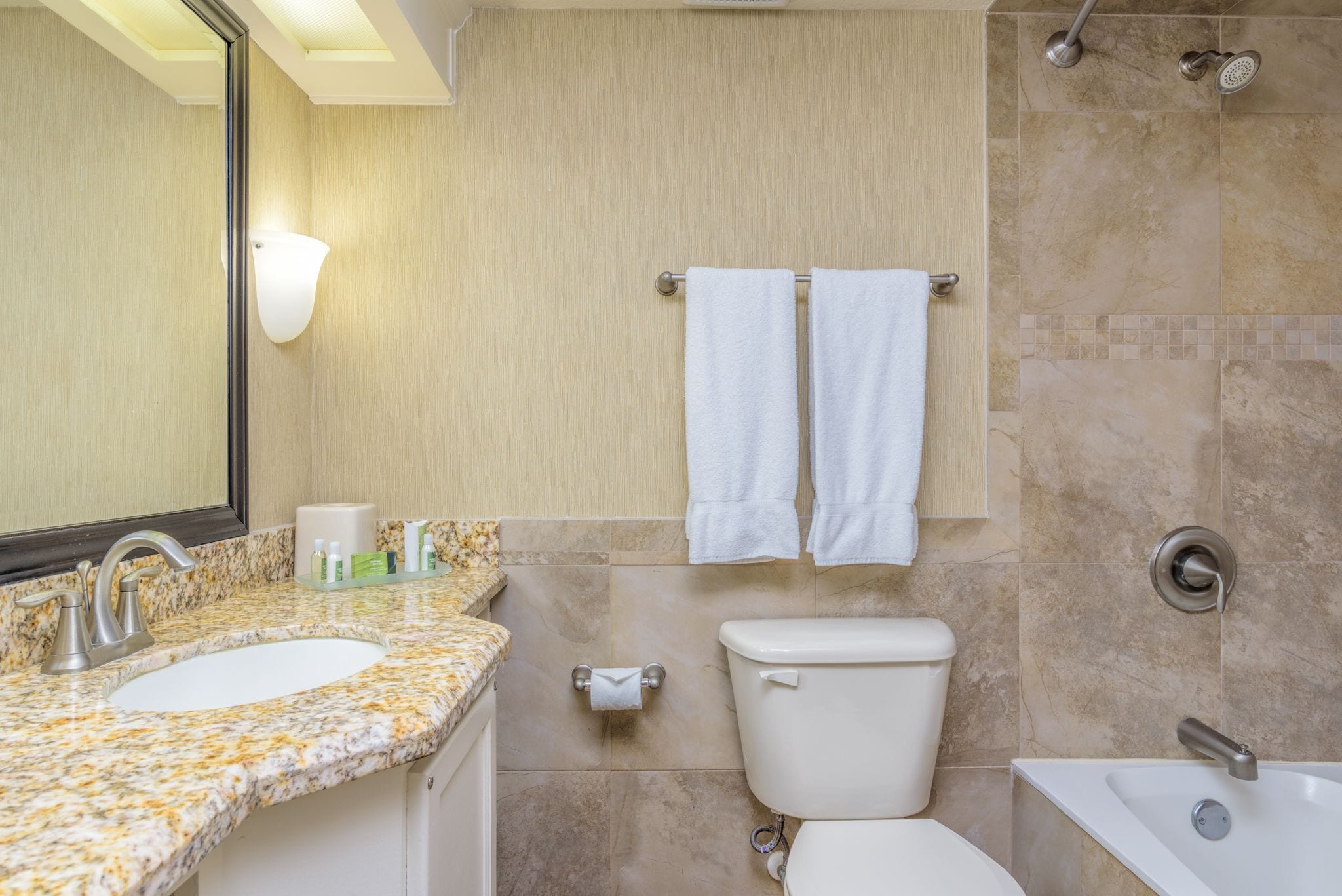 Une salle de bain équipée d’un lavabo avec un plan de travail en granit, de toilettes, d’une baignoire et d’un porte-serviettes avec deux serviettes blanches.