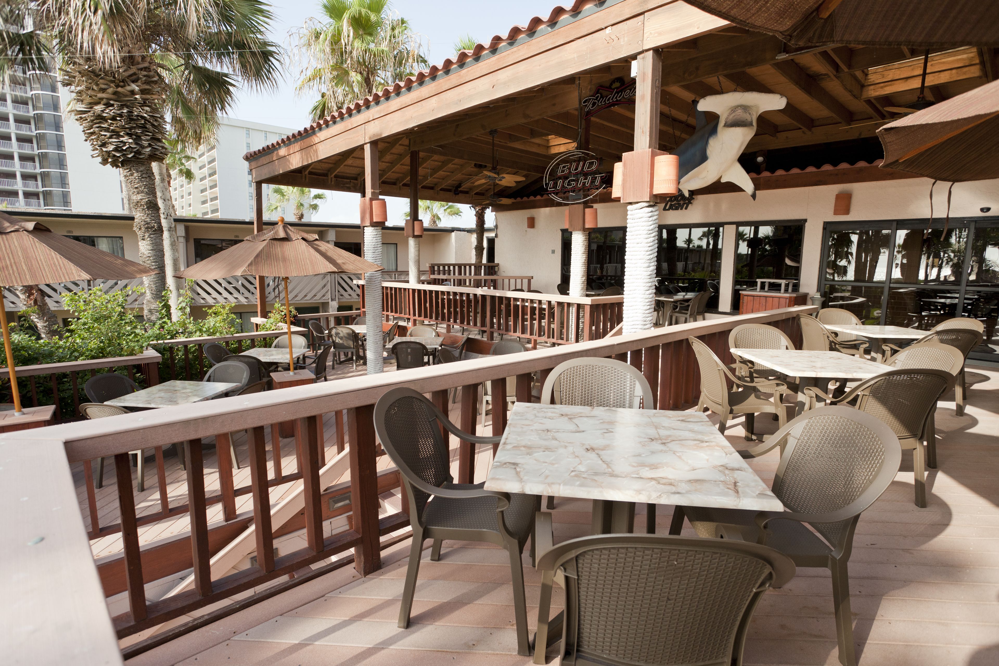 An outdoor patio area with tables and chairs, a wooden deck, and a view of palm trees and buildings in the background.