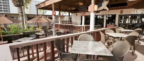 An outdoor patio area with tables and chairs, a wooden deck, and a view of palm trees and buildings in the background.