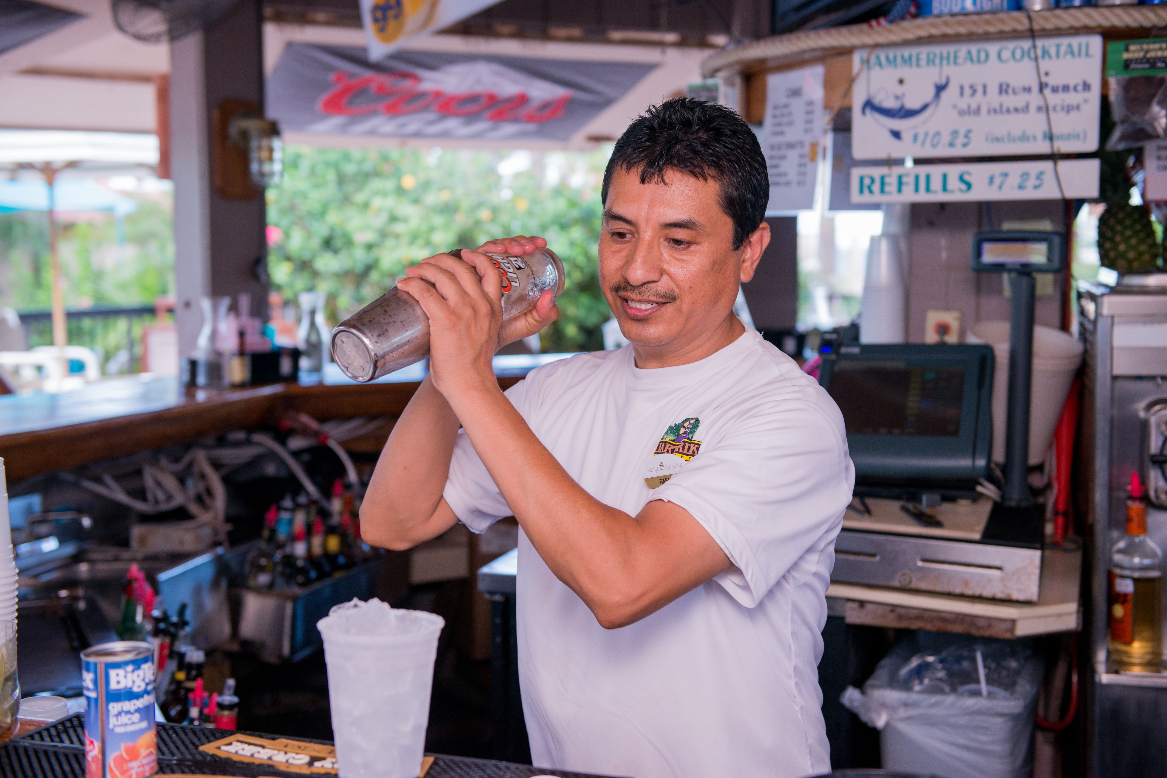 A bartender preparing a drink at a bar with a menu board in the background.