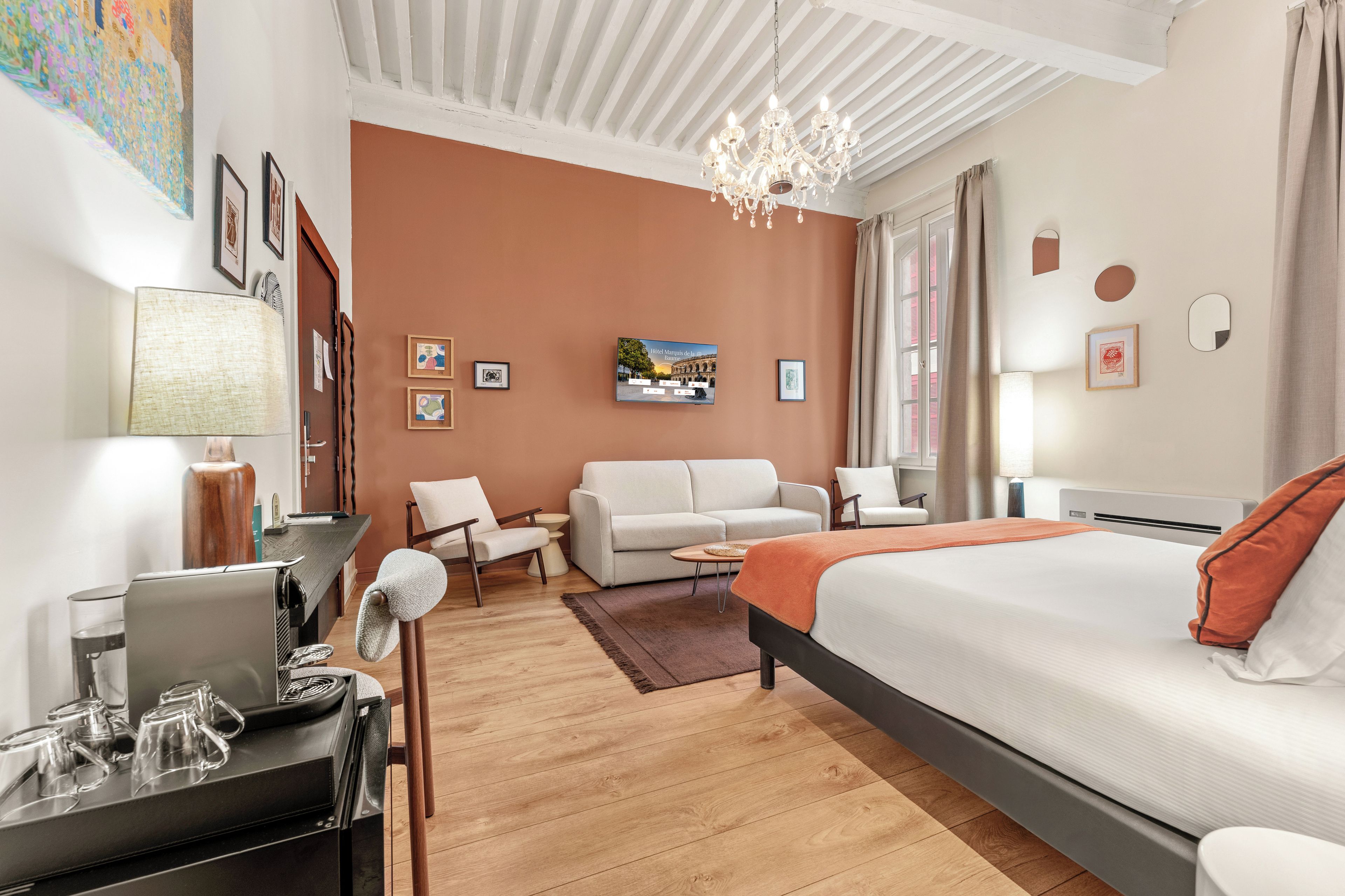 Ein Hotelzimmer mit einem Bett, einem Schreibtisch mit einem Laptop, einem Stuhl, einer Couch, einer Kristallleuchter und einem Bild an der Wand.