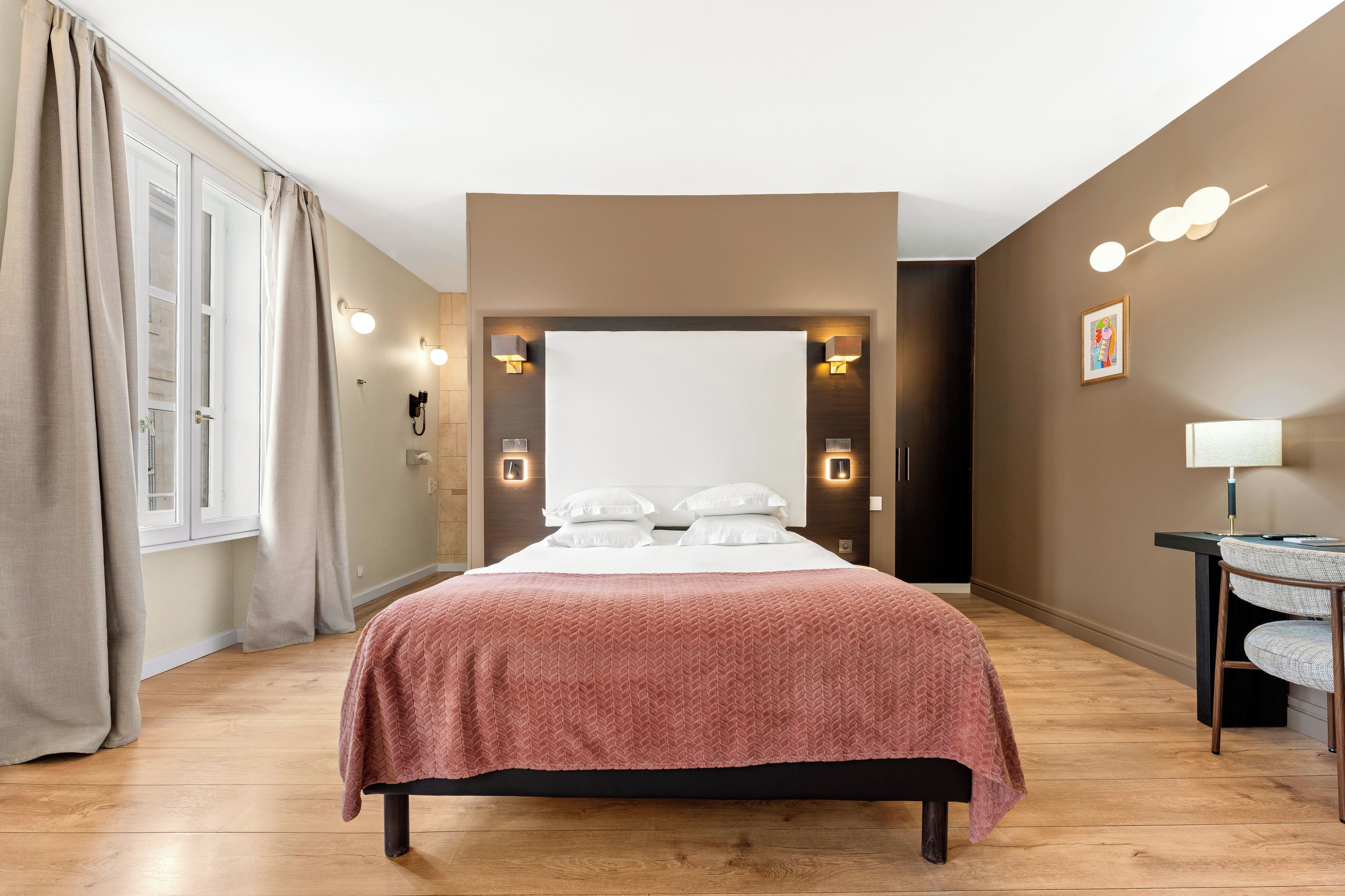 Een hotelkamer met een groot bed, een bureau met stoel, een raam met gordijnen en wandlampen.