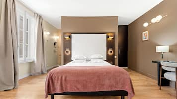 A hotel room with a large bed, a desk with a chair, a window with curtains, and wall-mounted lighting.
