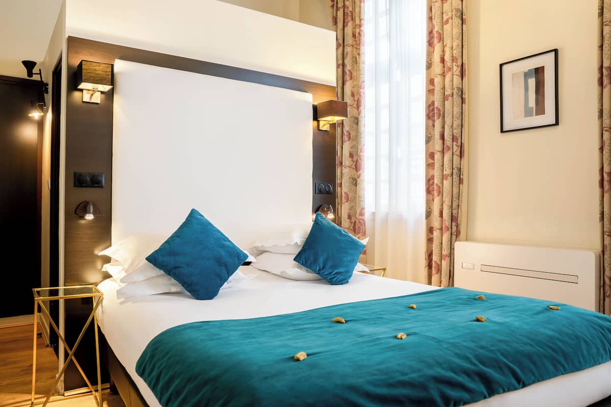 Un lit bien fait, avec une couvre-lit turquoise et deux oreillers bleus, un luminaire fixé au mur et un tableau encadré accroché au mur.