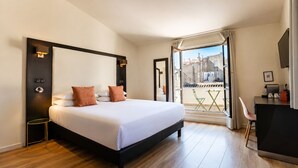 A hotel room with a large bed, a desk, and a balcony with a view of buildings.
