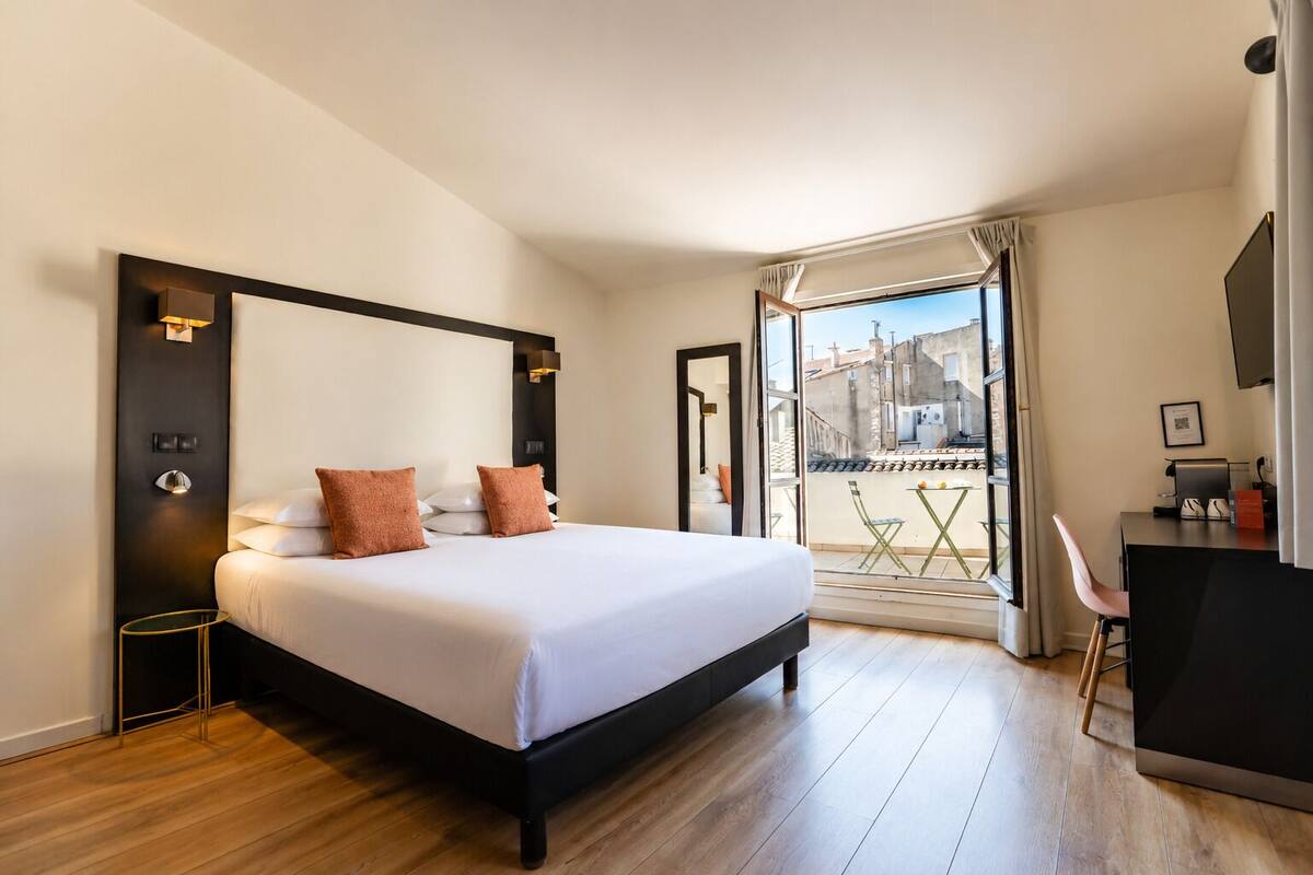 Une chambre d’hôtel avec un grand lit, un bureau et un balcon donnant sur les bâtiments.
