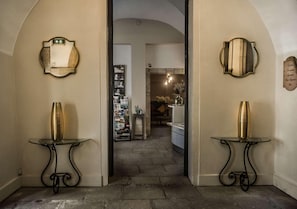 Un couloir avec deux tables en verre, chacune ornée d’un vase en laiton, mène à une pièce dotée d’une cheminée et d’un lustre.