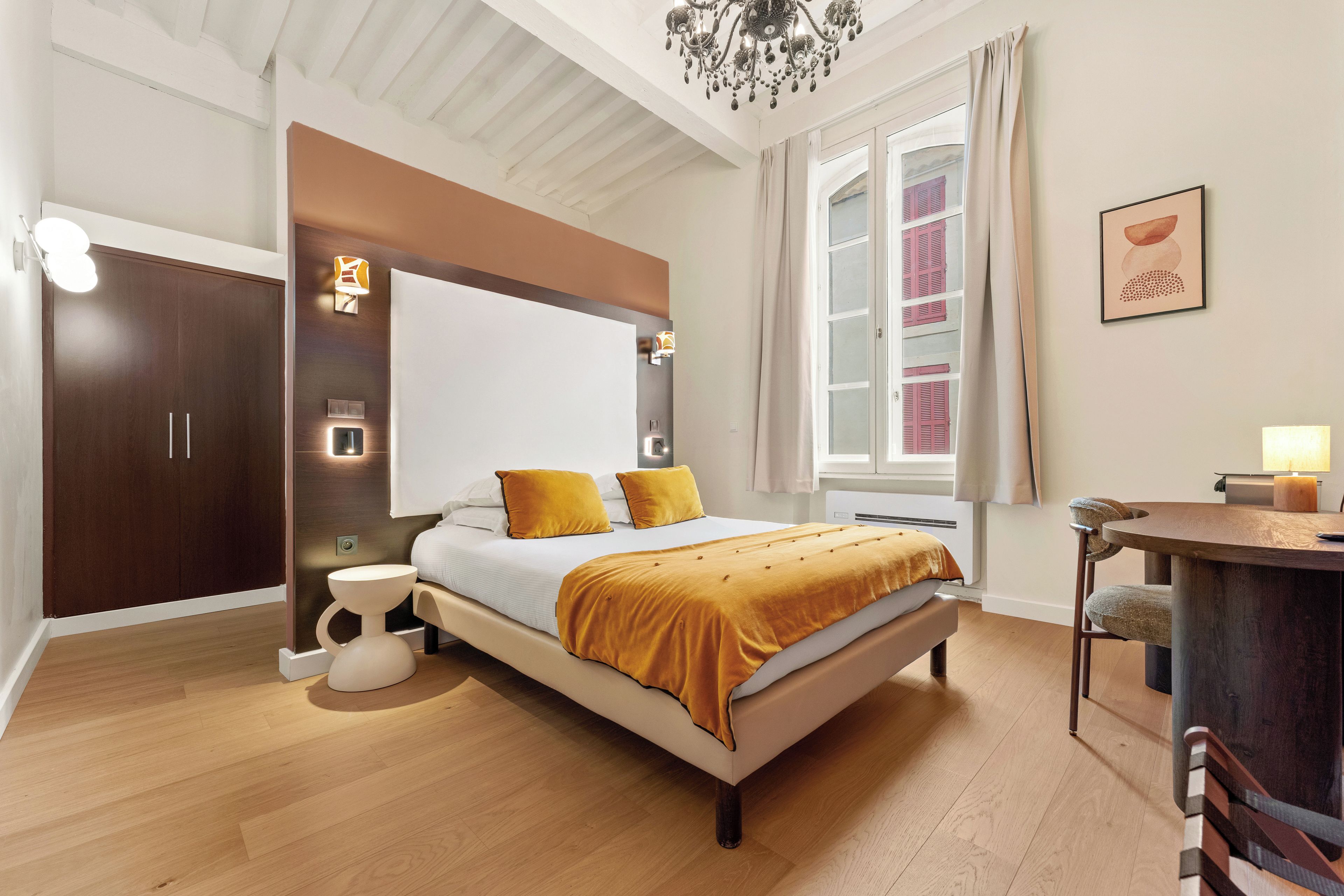 A modern bedroom with a large bed, bedside lamps, a desk, and a view of buildings through the window.