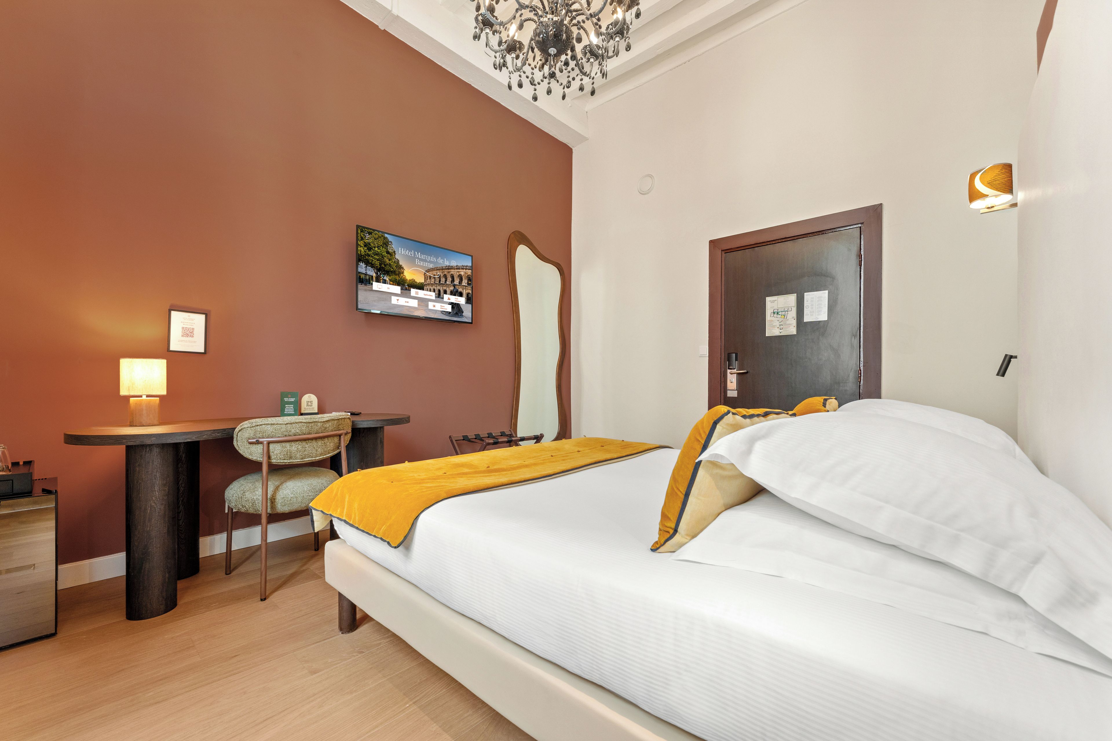 A hotel room with a bed, a desk, a chair, a mirror, and a chandelier.