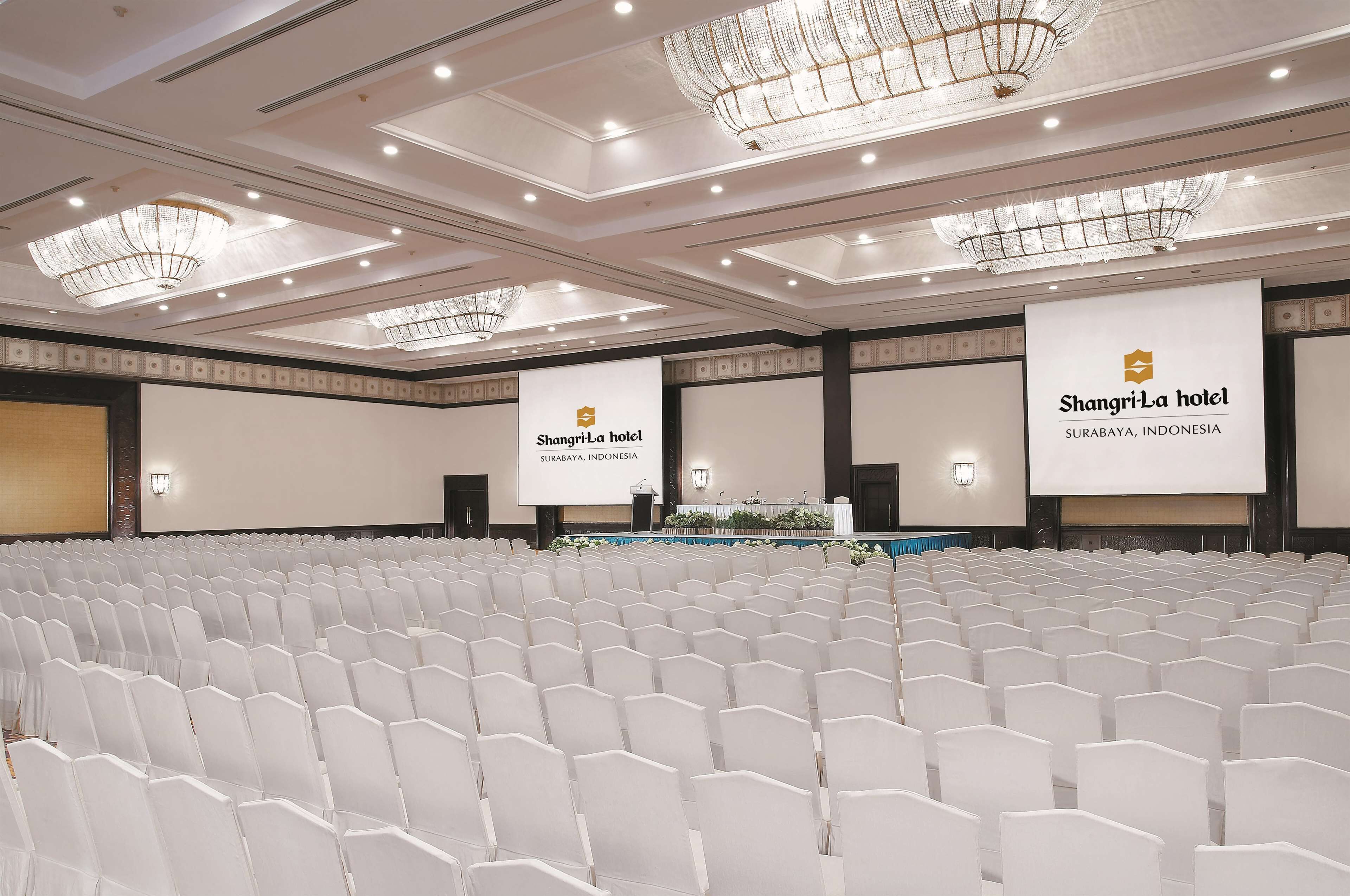 Een vergaderzaal met rijen witte stoelen, kroonluchters en een projectiescherm waarop de naam van het hotel staat.
