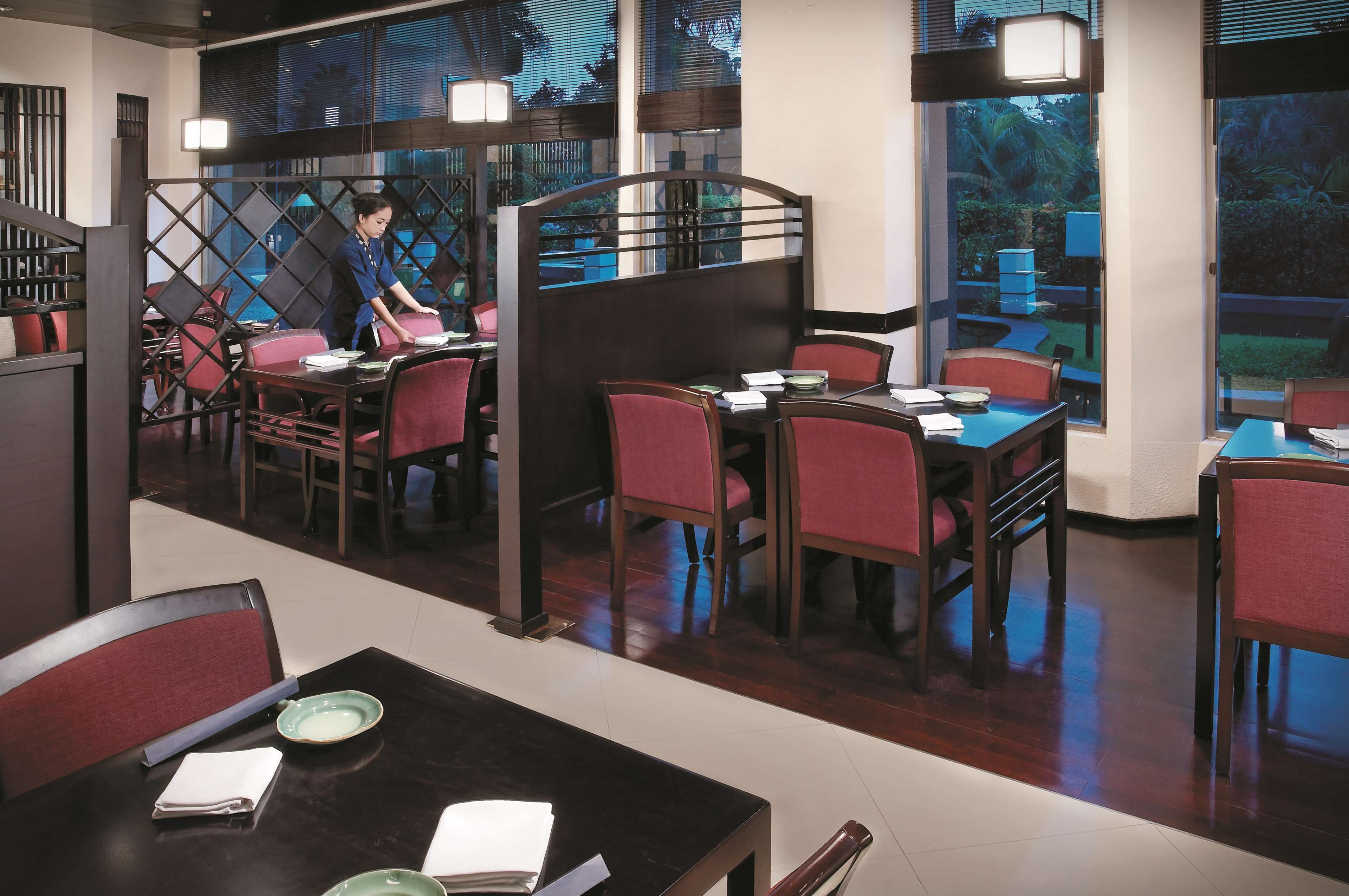 Un restaurant avec des tables en bois et des chaises rouges, un serveur qui prend les commandes à une table, et de grandes fenêtres donnant sur un paysage verdoyant.