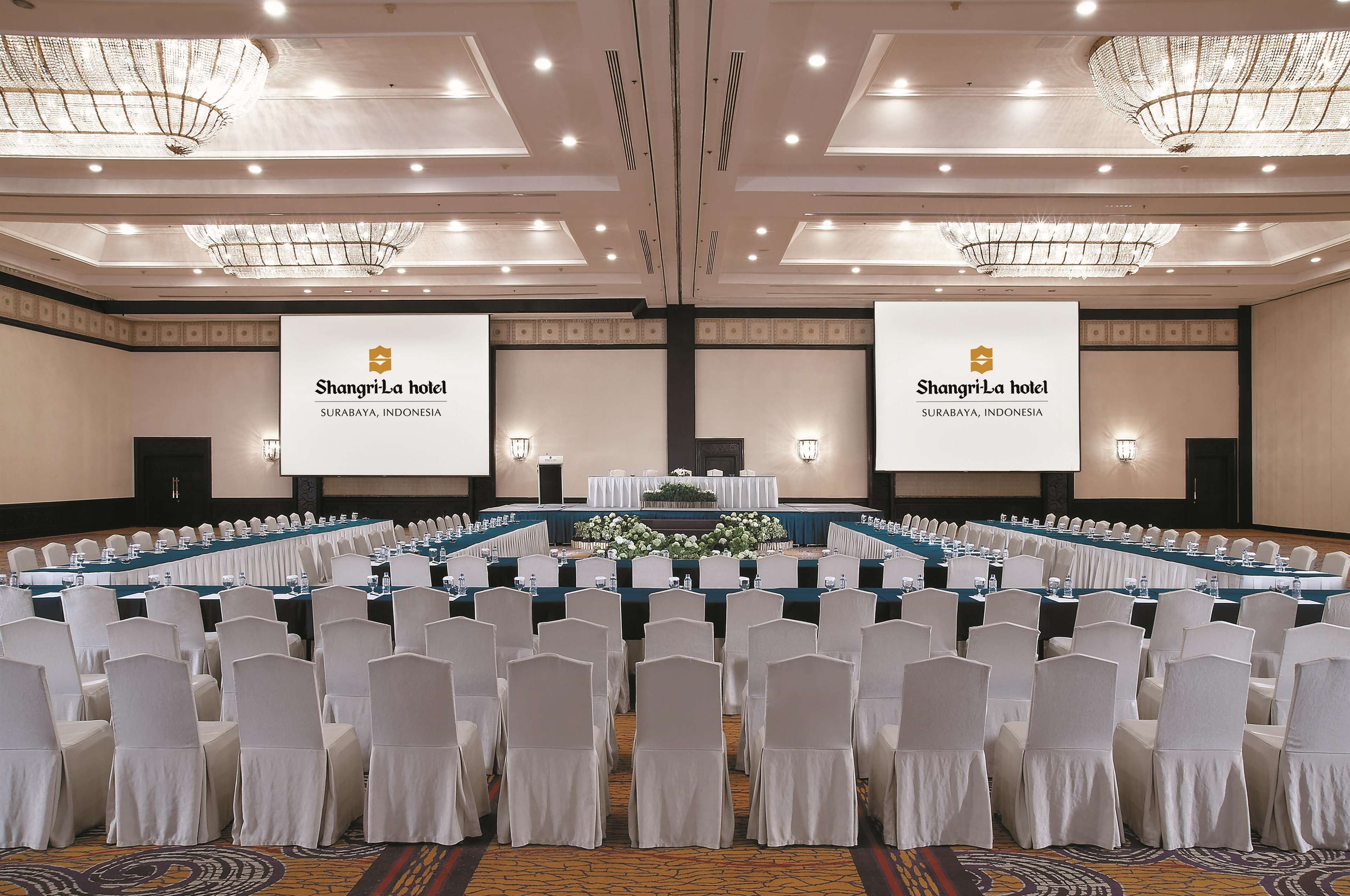A conference room at Shangri-La hotel in Surabaya, Indonesia, with rows of white chairs, a long table, and two large screens displaying the hotel name.