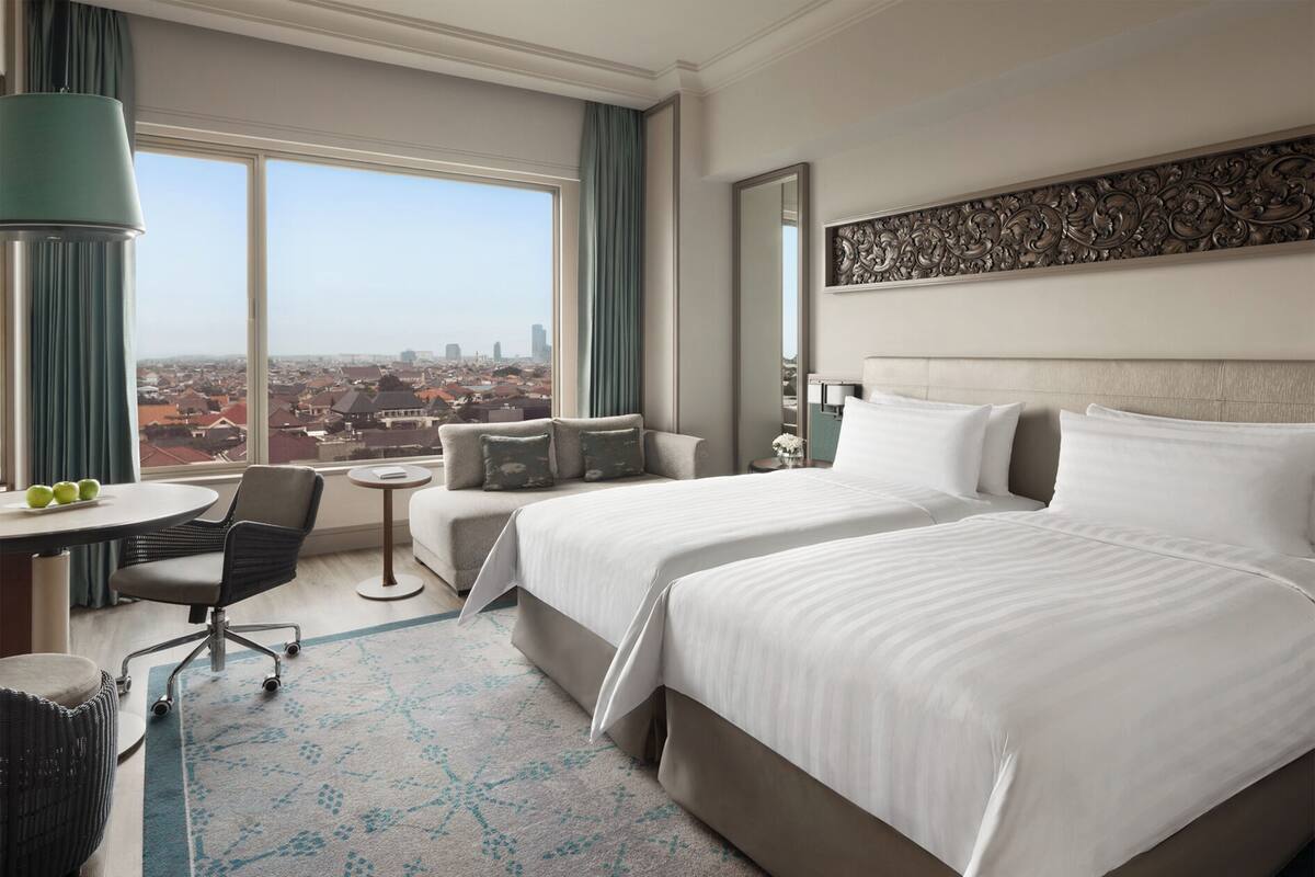 Een hotelkamer met twee bedden, een bureau, een stoel, een tafeltje en uitzicht op de stad.