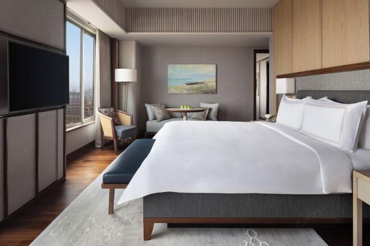 A modern hotel room with a large bed, a TV, a small table with chairs, and a painting on the wall.
