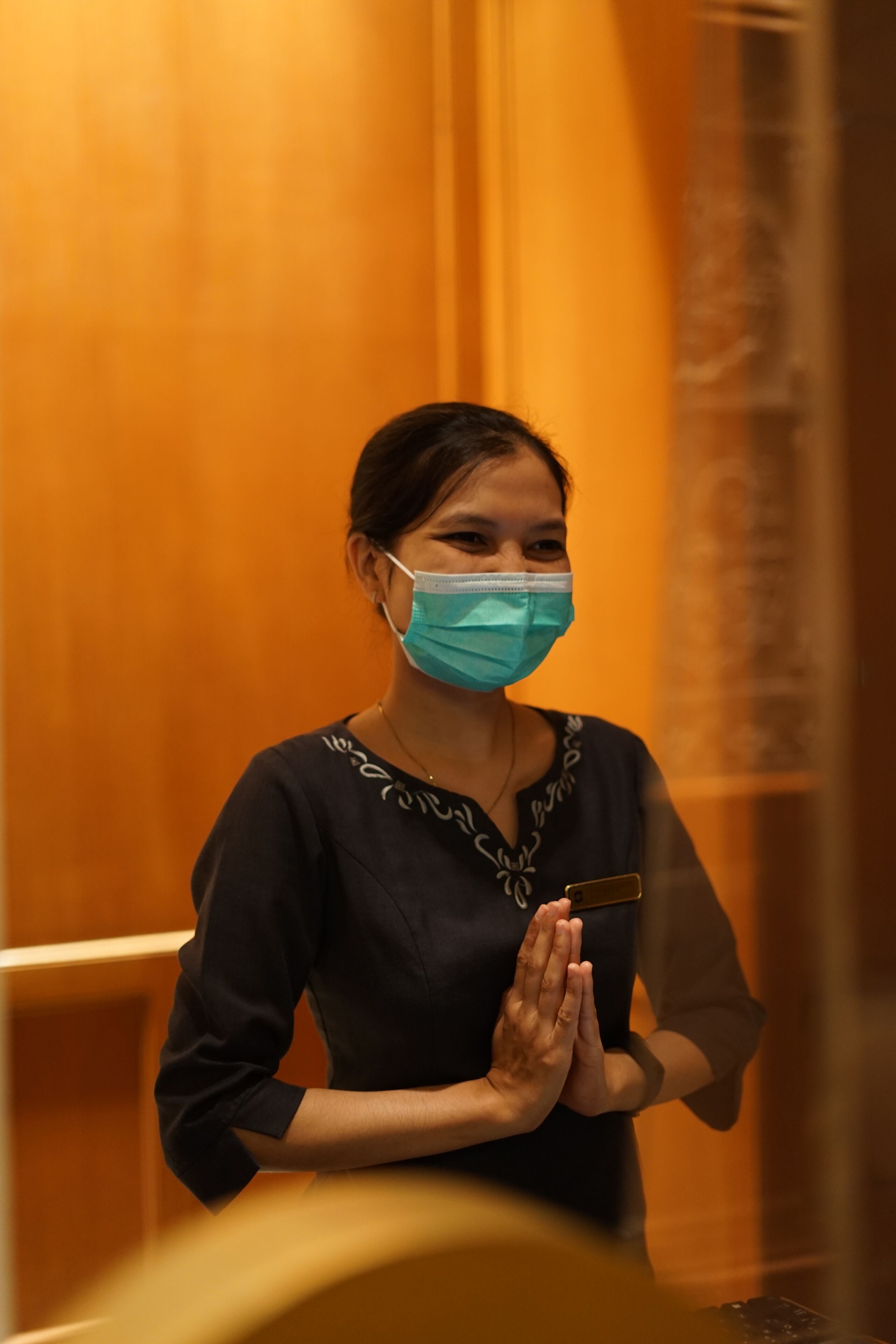A person standing in an elevator with a name tag, wearing a dark top with a pattern, and a face mask.