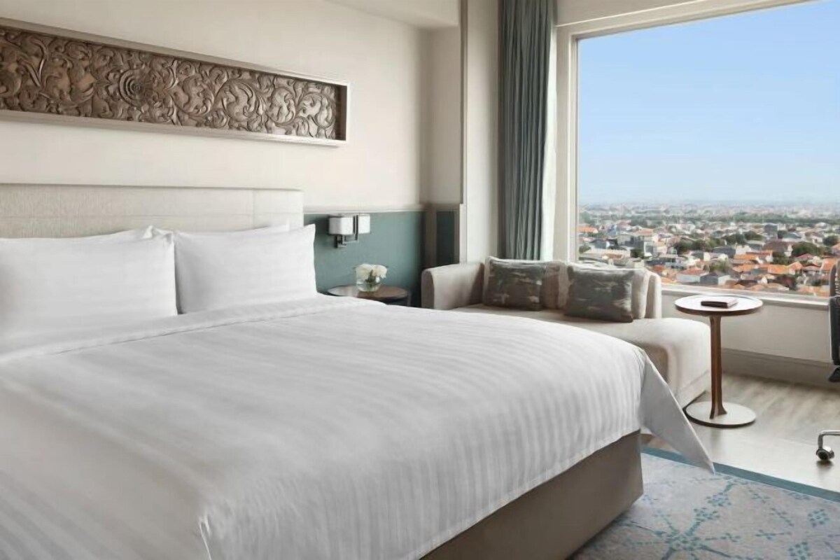 A hotel room with a large bed, a view of the city, a small table, and a decorative wall piece.