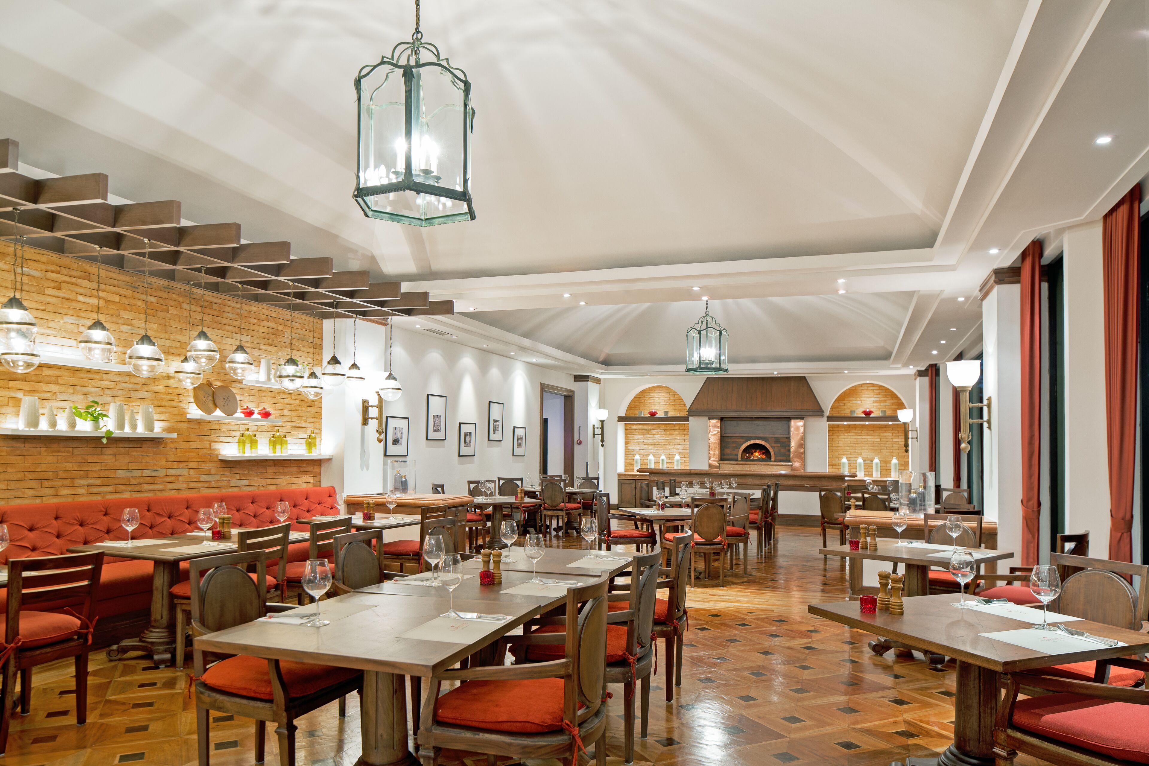 A spacious restaurant with wooden flooring, red cushioned booths, and hanging pendant lights. Tables are set with wine glasses and menus.