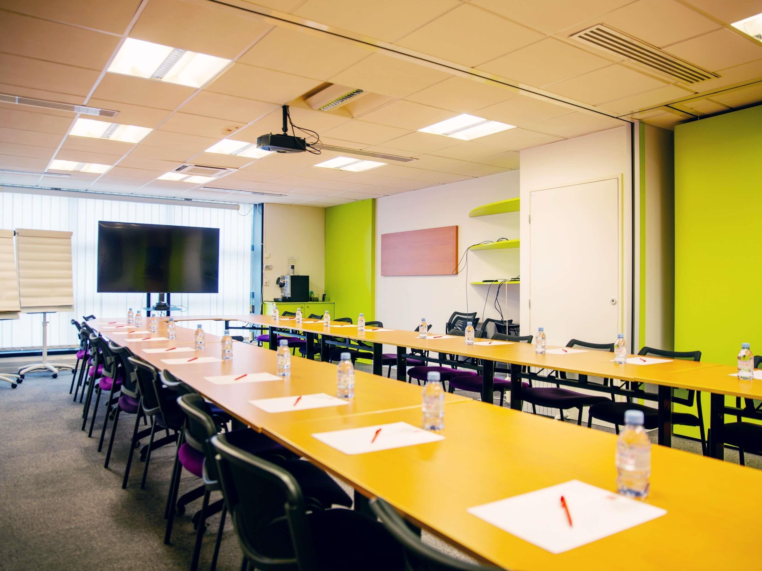 A conference room with long tables, chairs, a projector, and a large screen.