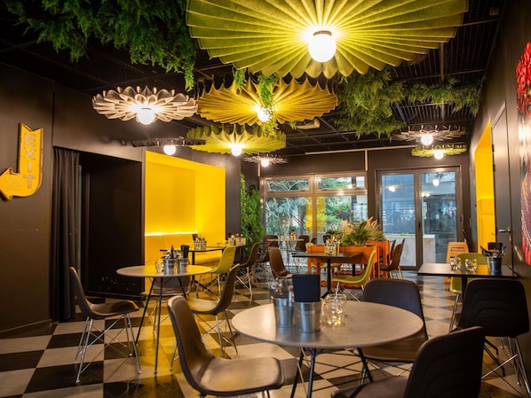 Een restaurant met een geruit vloertje, ronde tafels en stoelen. Er hangen lampen en begroeiing aan het plafond.