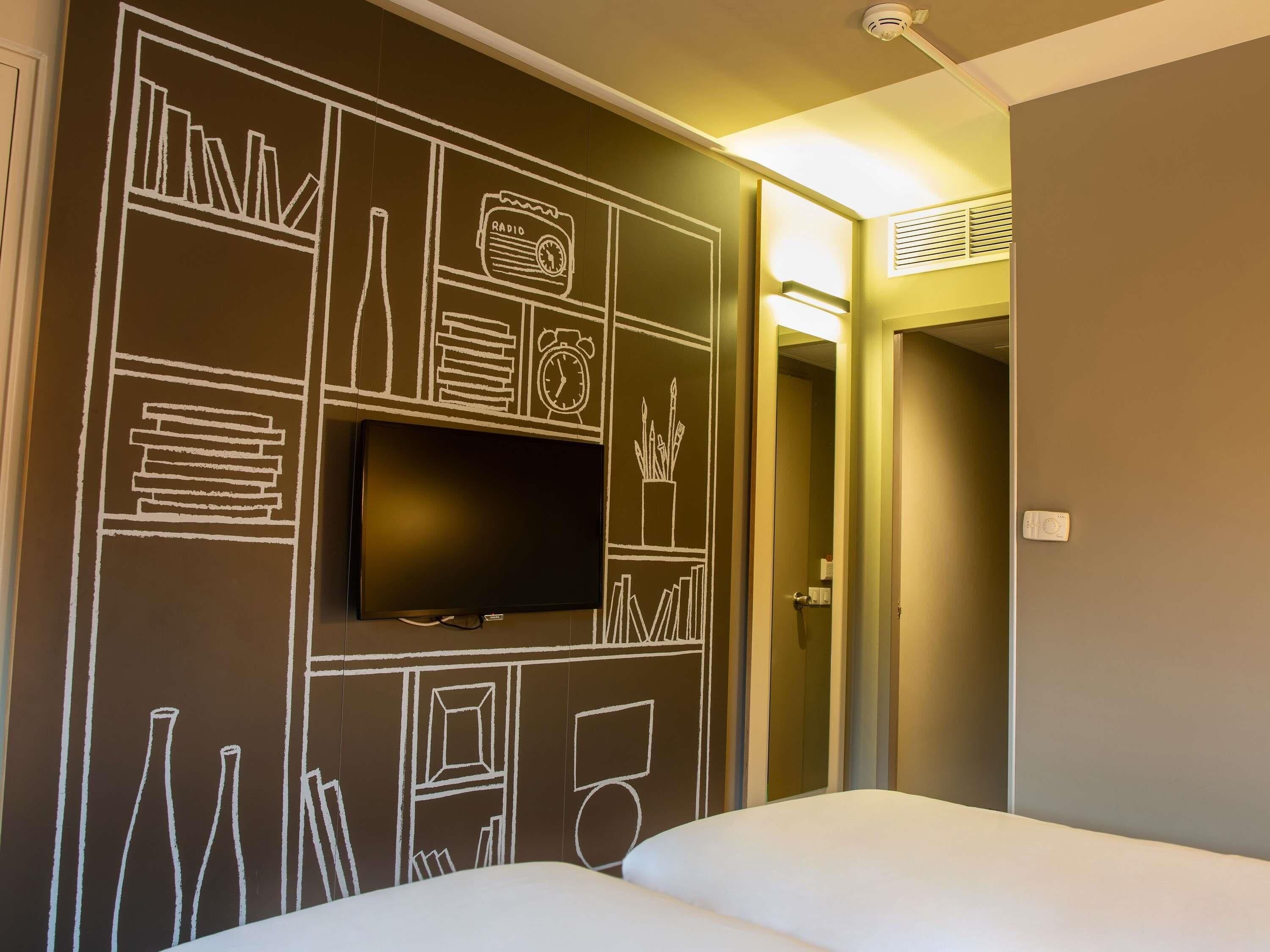 A hotel room with a large flat-screen TV mounted on the wall, a bed with white bedding, and a door leading to another room.