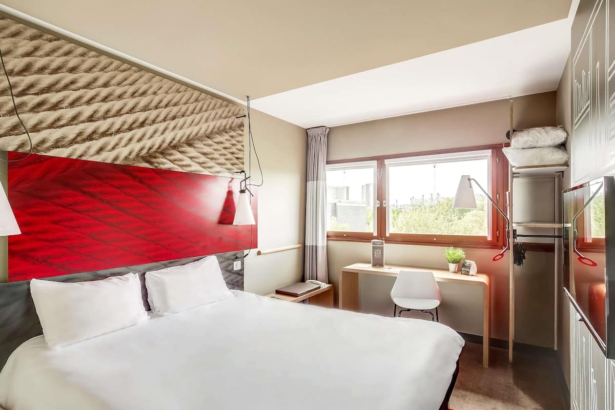 Une chambre d’hôtel avec un lit, un bureau, une chaise, une fenêtre donnant sur un paysage agréable et un mur d’accent rouge.