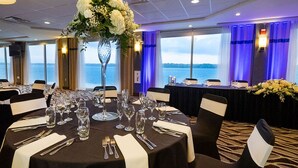 Un salón de banquetes con mesas redondas dispuestas para un evento formal, ventanas amplias con vista al agua y un jarrón decorativo con flores.