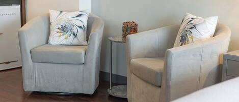Deux fauteuils ornés de coussins à motifs floraux, une petite table d’appoint sur laquelle est posé un vase décoratif, et un plancher en bois.
