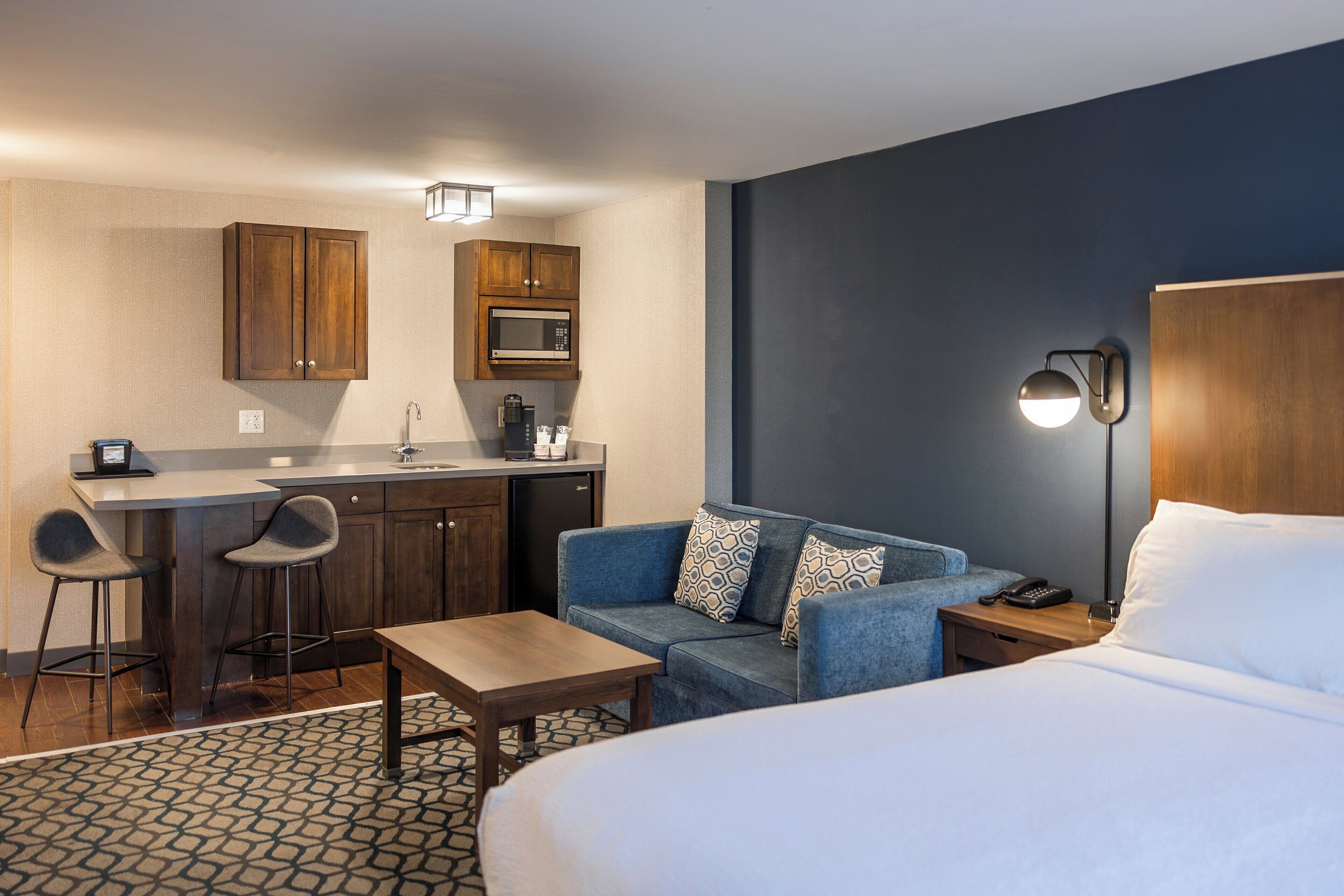 A hotel room with a bed, a sofa, a small table, a kitchenette with a microwave, and a bar area with stools.