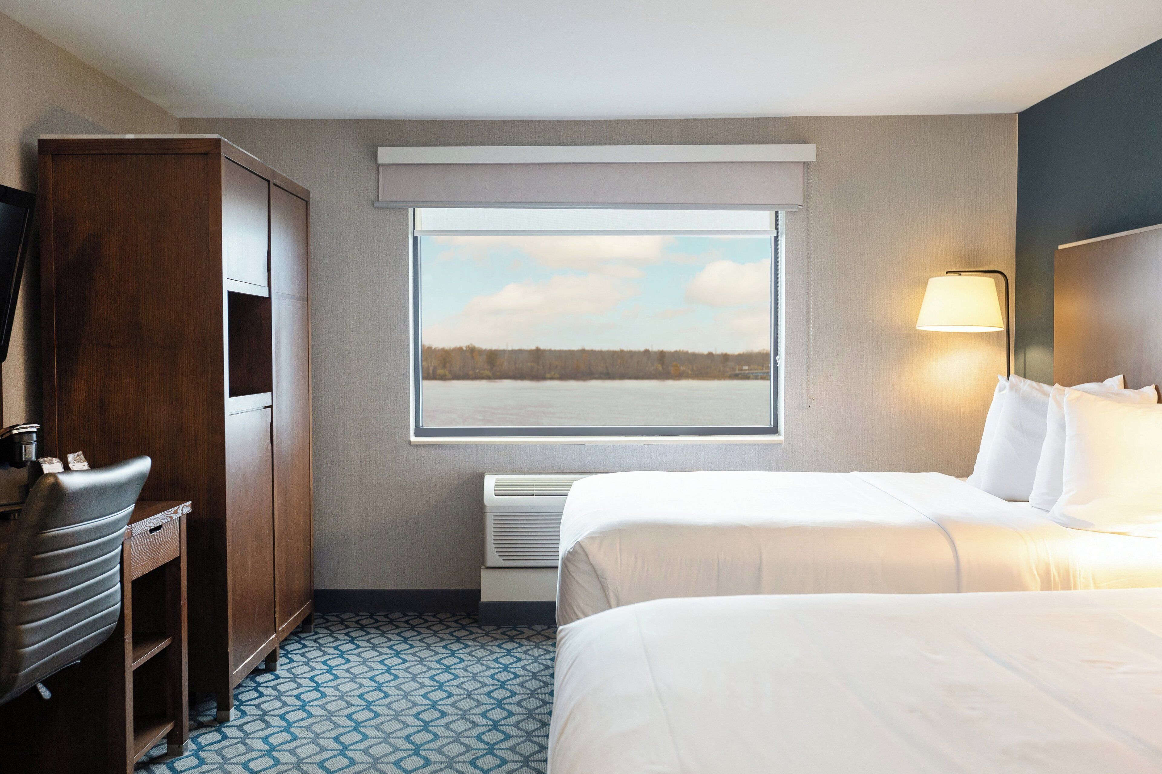 A hotel room with a bed, a desk, a wardrobe, a window with a view of trees, and a wall-mounted lamp.
