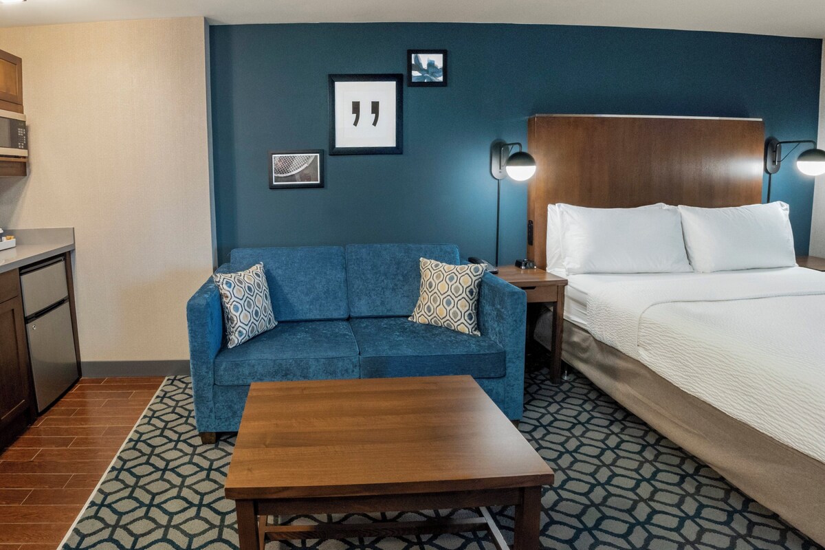 A hotel room with a blue sofa, a wooden table, a bed with white linens, a nightstand with a lamp, and a kitchenette with a microwave.