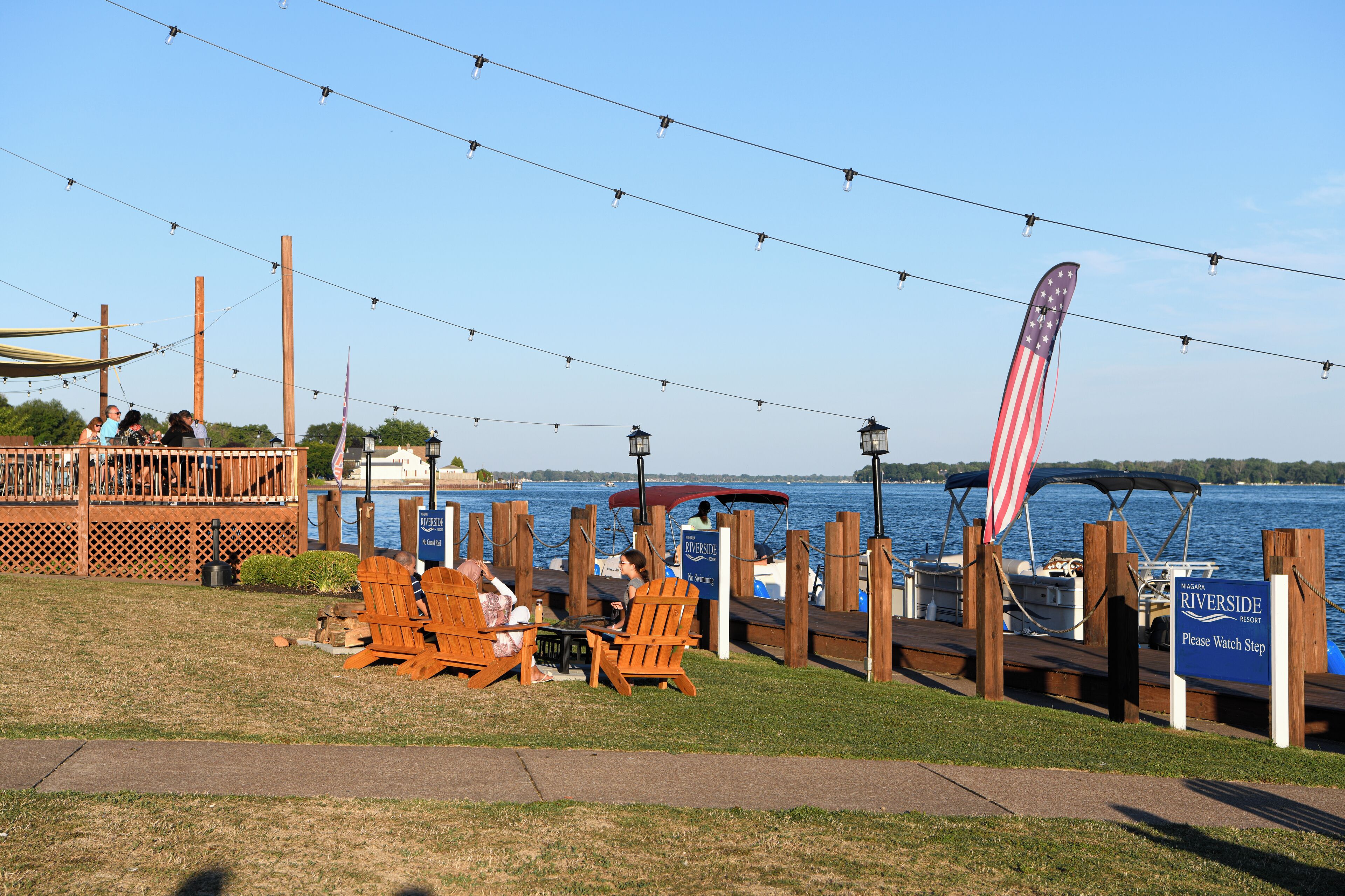 A lakeside deck with wooden chairs, a dock with boats, and a sign for Riverside Resort.