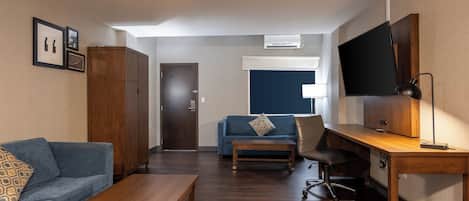 Habitación de hotel con un escritorio de madera, un sofá azul, una mesa baja de madera, un televisor y un equipo de aire acondicionado.