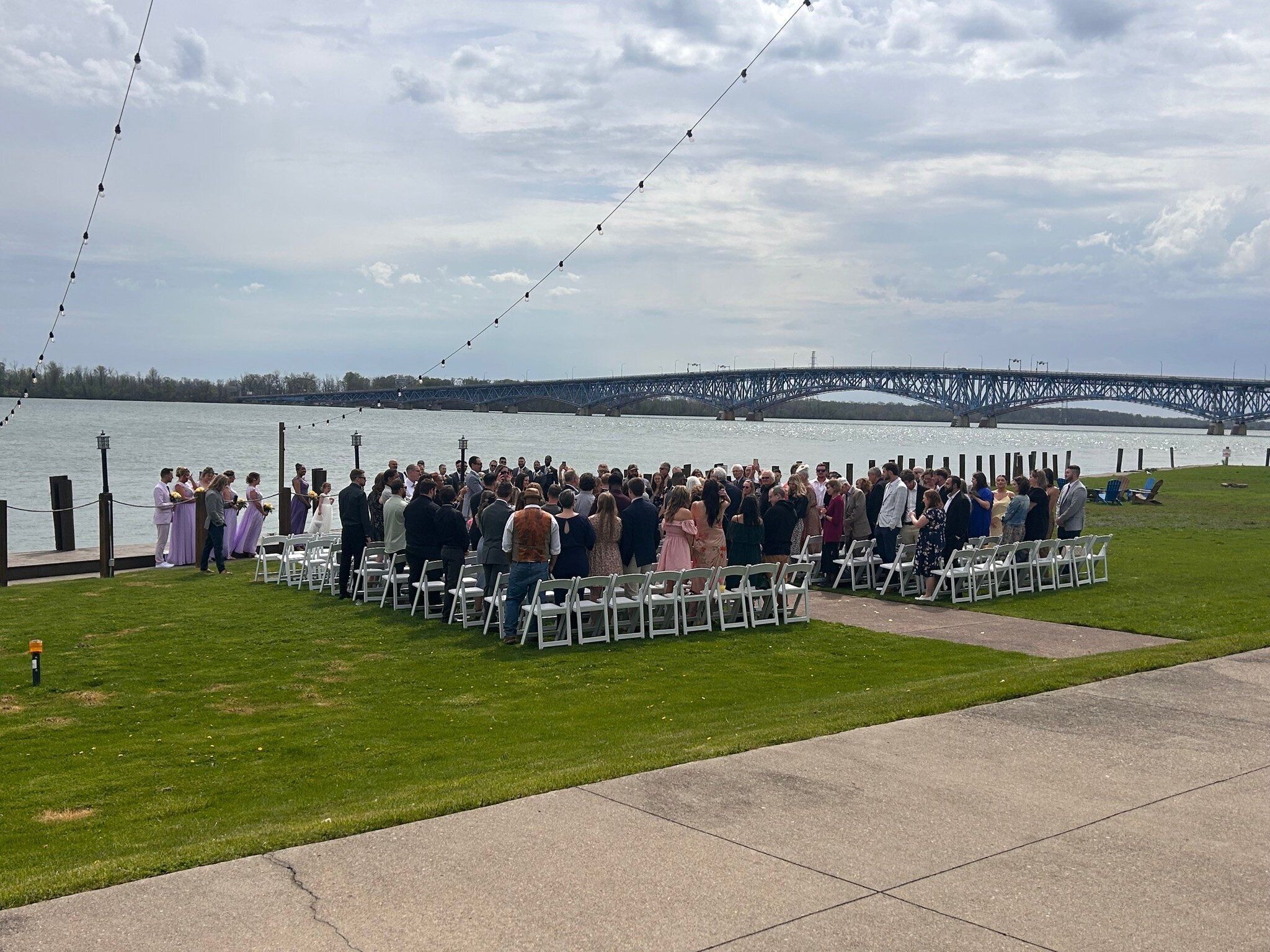 A wedding ceremony outdoors with guests seated in rows facing a body of water and a bridge in the background.
