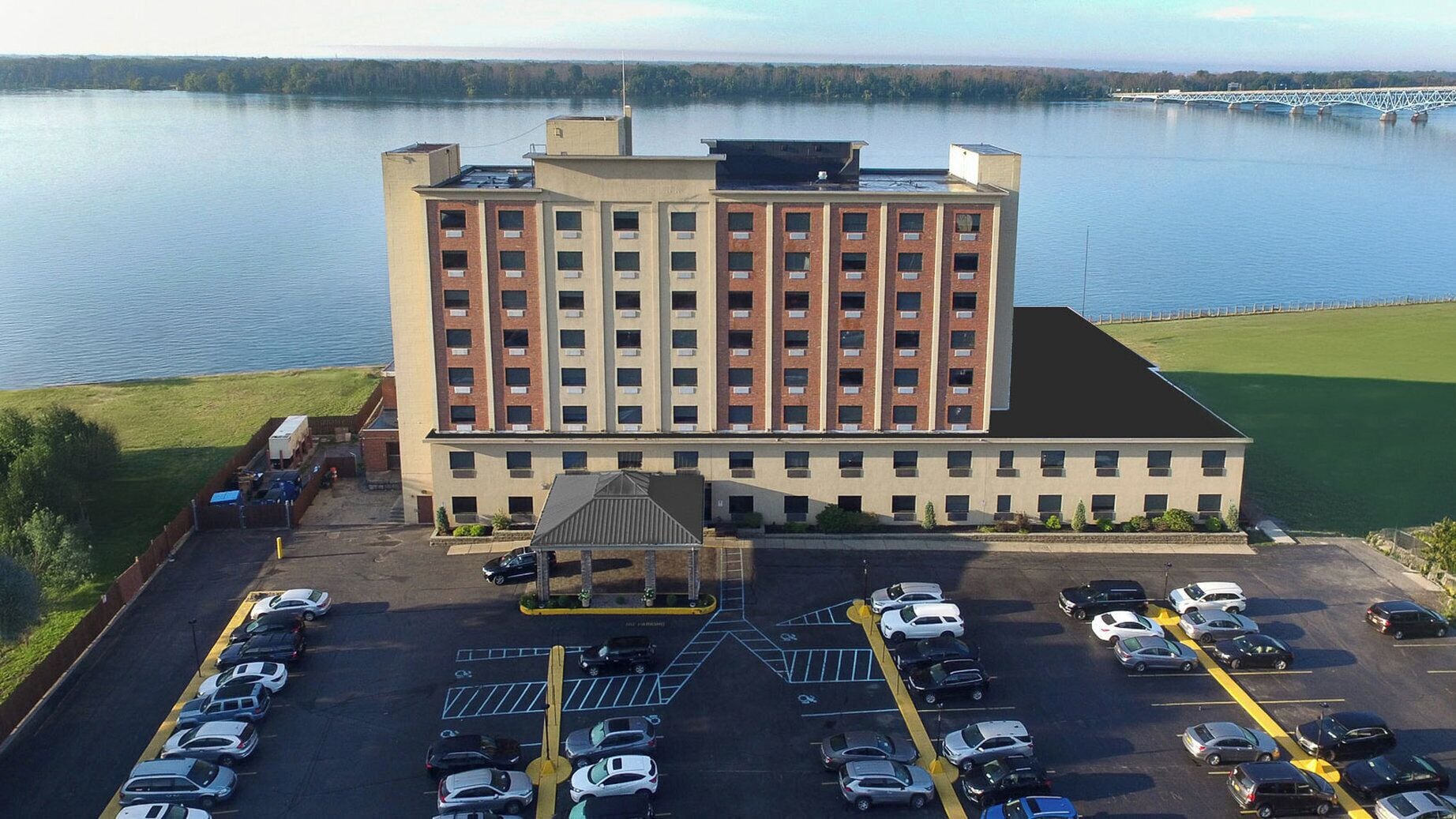 A multi-story hotel with a parking lot in front, a small covered area, and a view of a lake.