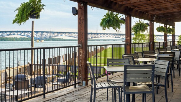 Un patio al aire libre con mesas y sillas, una pérgola de madera, plantas colgantes, vistas de un puente y una barandilla con vista al agua.