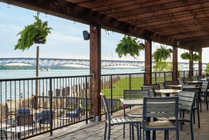 Eine Außenpatio mit Tischen und Stühlen, einer Holzpergola, hängenden Pflanzen, Blick auf eine Brücke und einem Geländer mit Blick auf ein Gewässer.