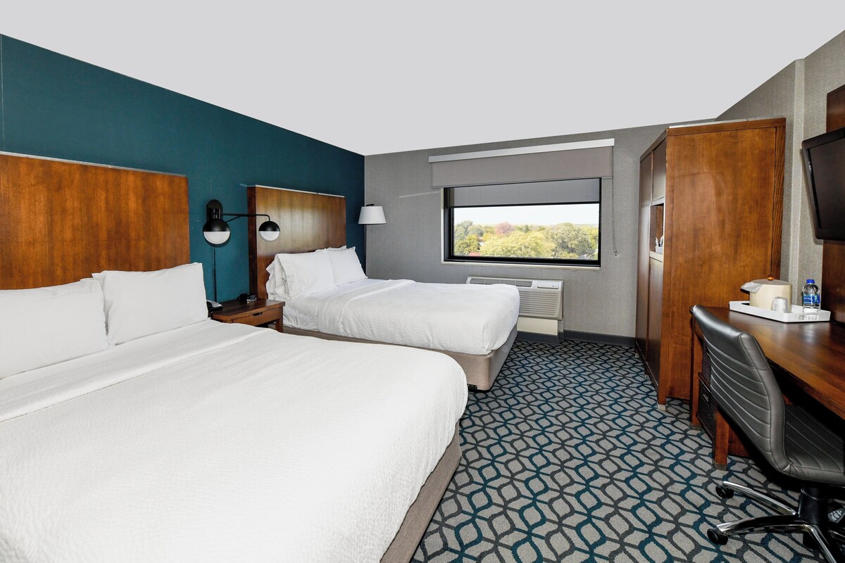 A hotel room with two beds, a desk, a chair, a TV, and a window with a view.
