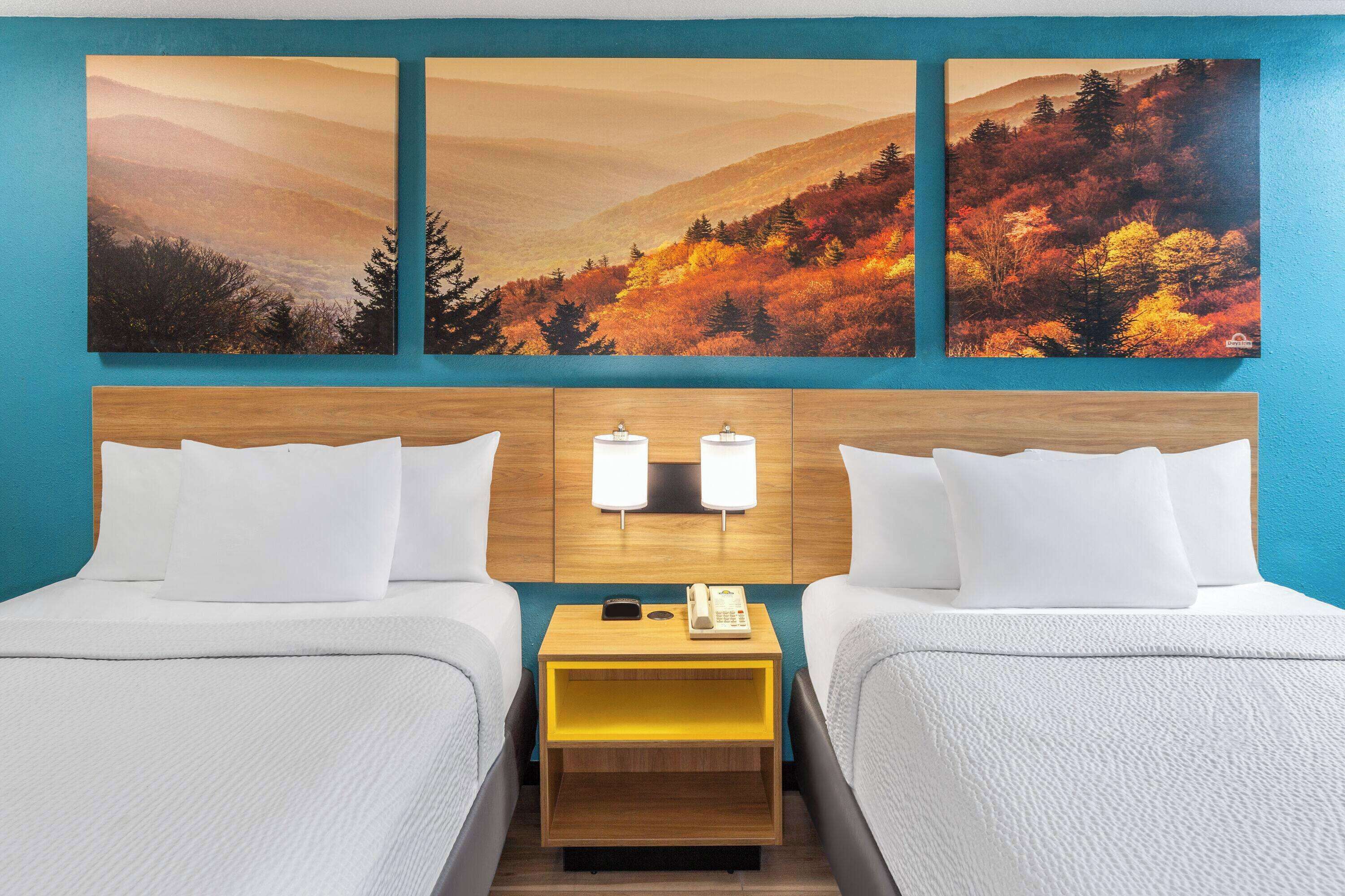 Habitación de hotel con dos camas, cabecera de madera, mesita de noche con teléfono, y pared decorada con tres pinturas de paisajes otoñales.