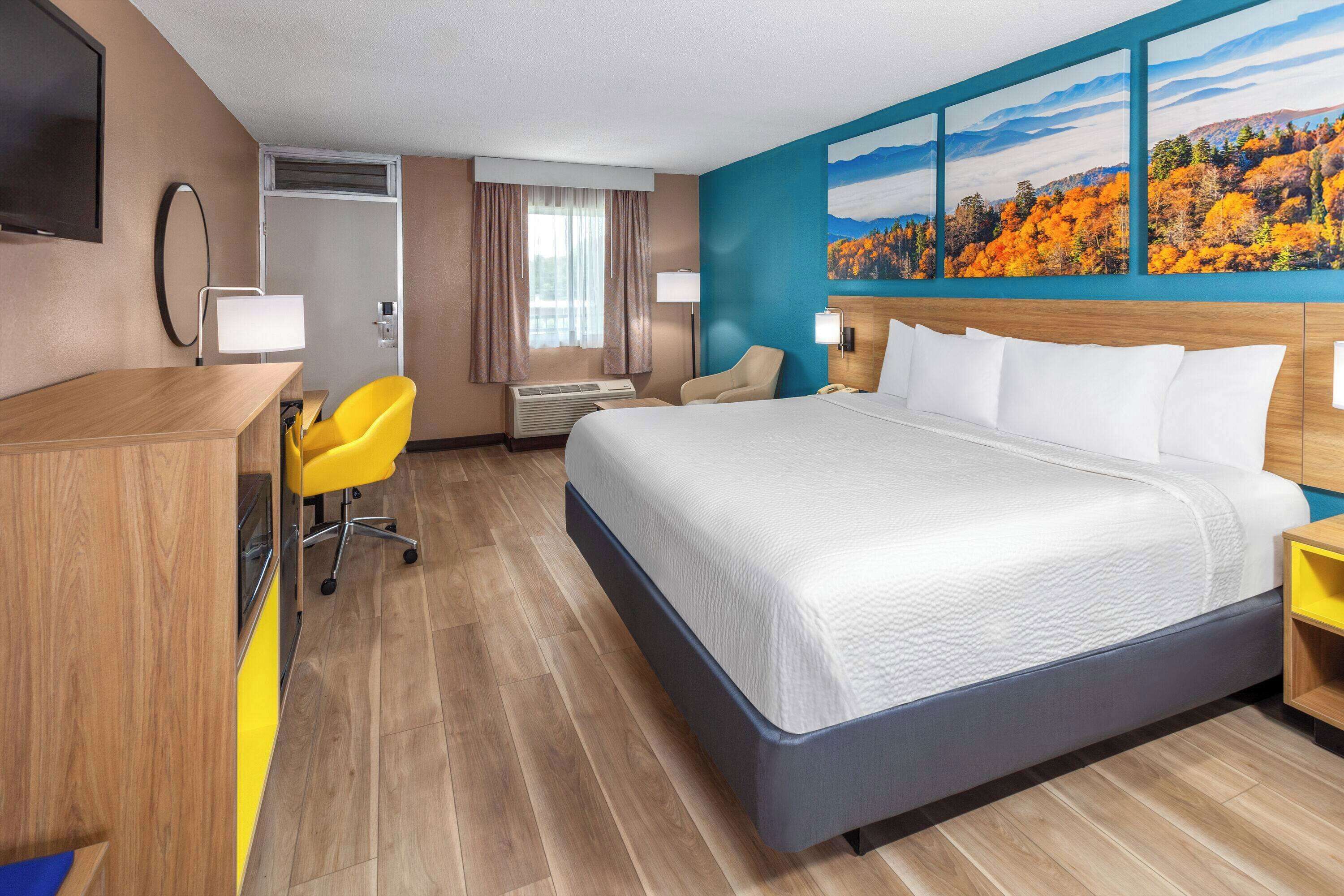 A hotel room with a large bed, a desk with a chair, an air conditioning unit, and a painting of autumn foliage on the wall.