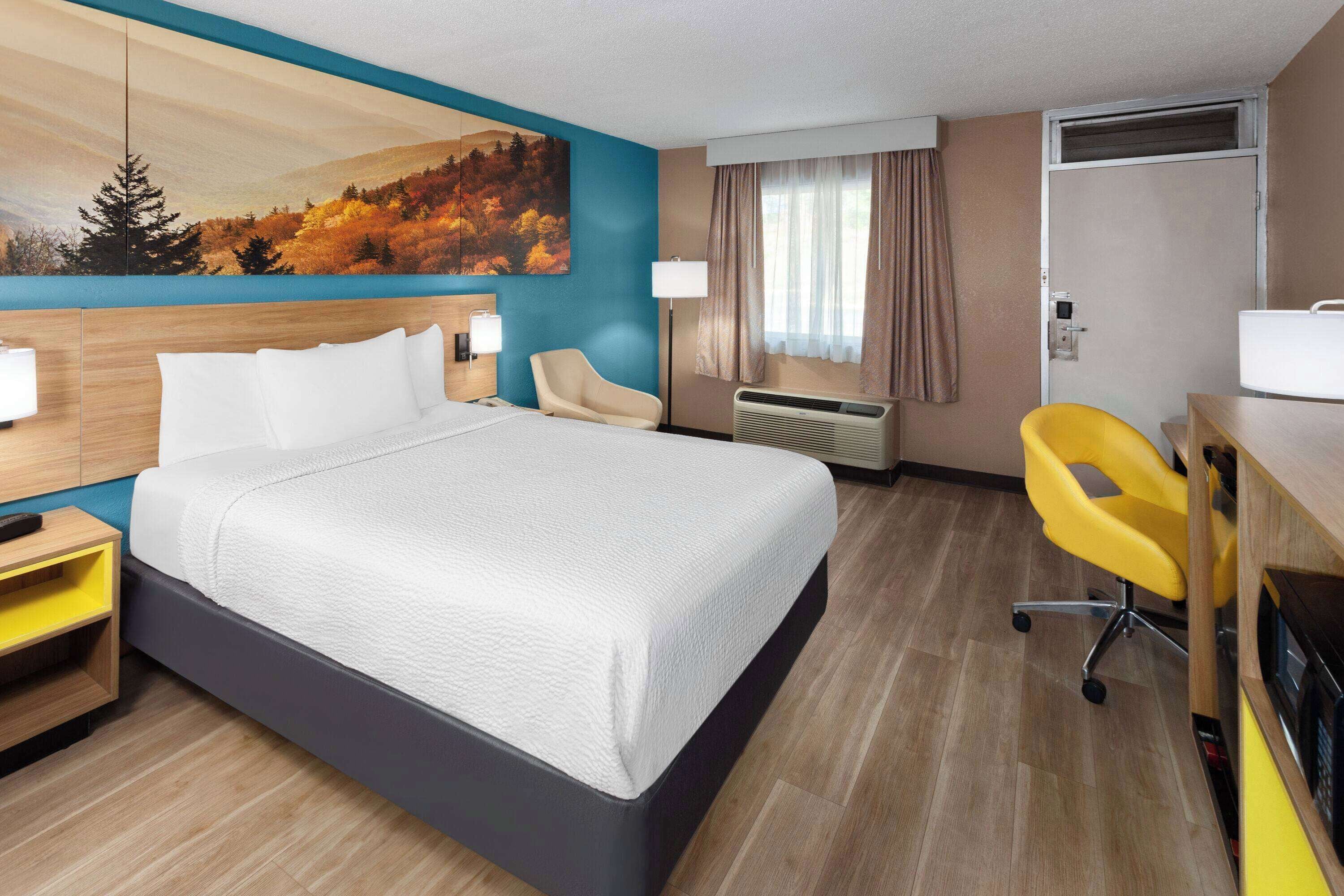 A hotel room with a large bed, a desk with a chair, a nightstand, a painting on the wall, and an air conditioning unit.
