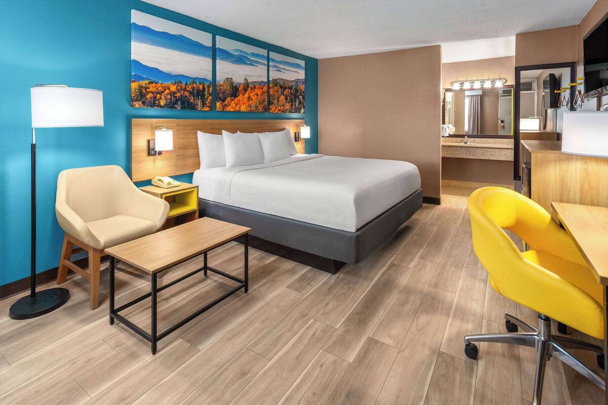Habitación de hotel con cama, escritorio, silla, mesita de noche, lámpara y un cuadro con follaje otoñal.