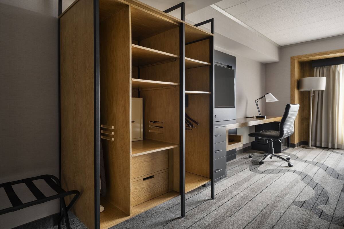 A modern hotel room with a wooden wardrobe, a desk with a chair, and a lamp.