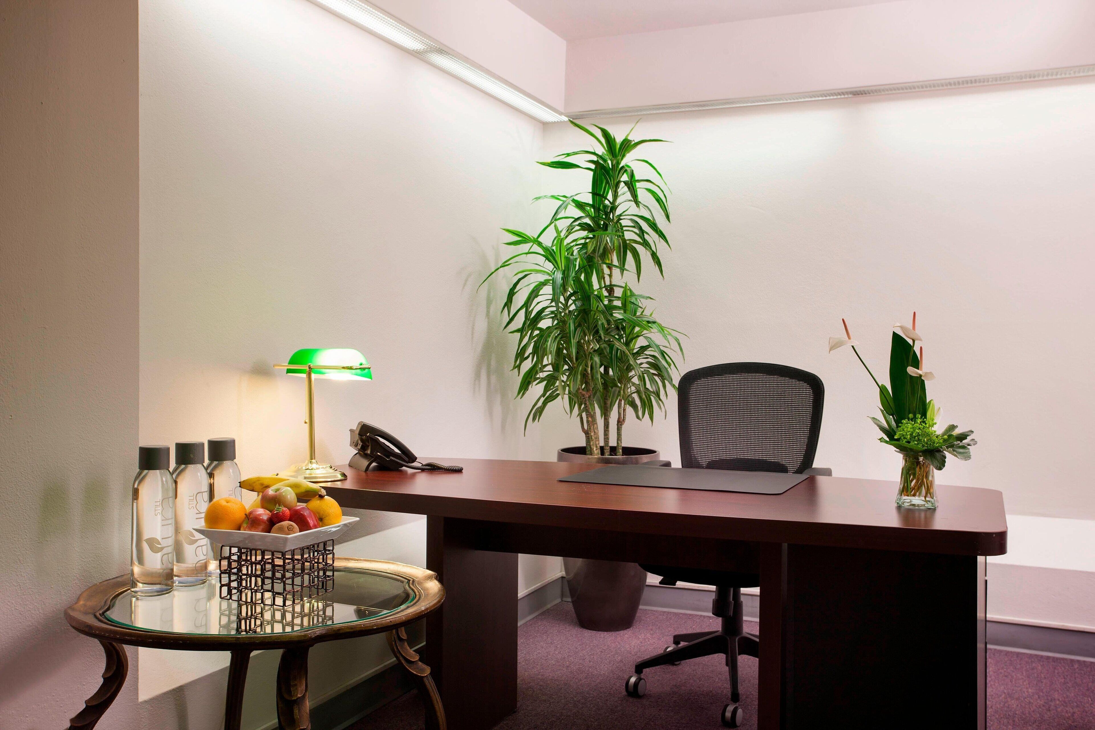 A small office with a desk, chair, potted plants, a lamp, and a fruit bowl.