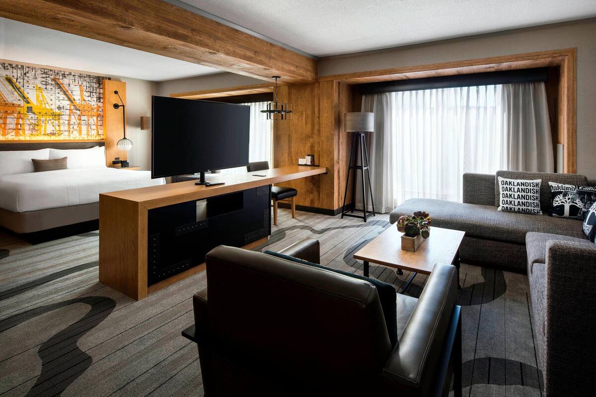 A hotel room with a bed, a TV, a desk, a sofa, and a large window with curtains.