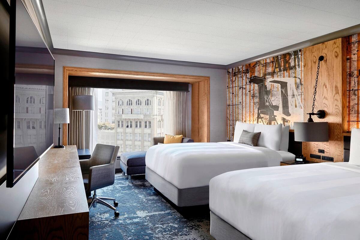 A hotel room with two beds, a desk, a chair, a large window with a view of a cityscape, and a wooden accent wall.