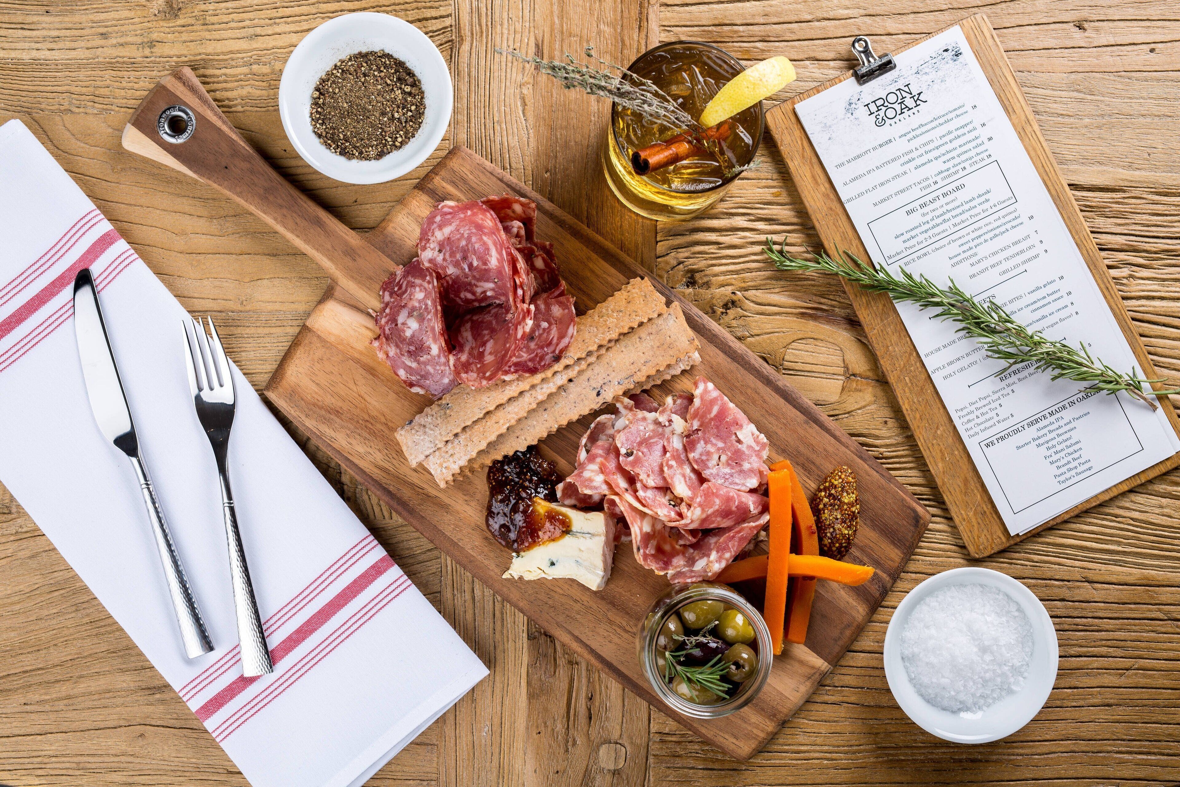 A wooden board with various cheeses, cured meats, and crackers, accompanied by a bottle of olive oil, a sprig of rosemary, and a menu on a clipboard.