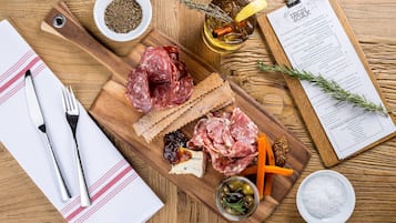 A wooden board with various cheeses, cured meats, and crackers, accompanied by a bottle of olive oil, a sprig of rosemary, and a menu on a clipboard.